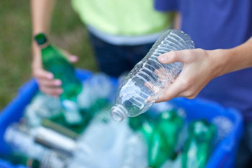 Close-up of a person's hand holding a clear plastic bottle with a ridged texture, preparing for rubbish collection outdoors. In the background, another individual wearing a bright yellow shirt and a green wristband is seen near a blue recycling bin filled with various plastic bottles, containers, and packaging materials. The scene is set on a paved or grassy area, indicating an environment where waste and recycling are being sorted or collected, consistent with rubbish removal services in Tower Hamlets. The focus on the plastic waste and the outdoor setting highlights the importance of proper disposal and recycling efforts carried out by companies like Garden Clearance Tower Hamlets. Natural daylight illuminates the scene, emphasizing the different shades of green and transparent plastics among the waste.