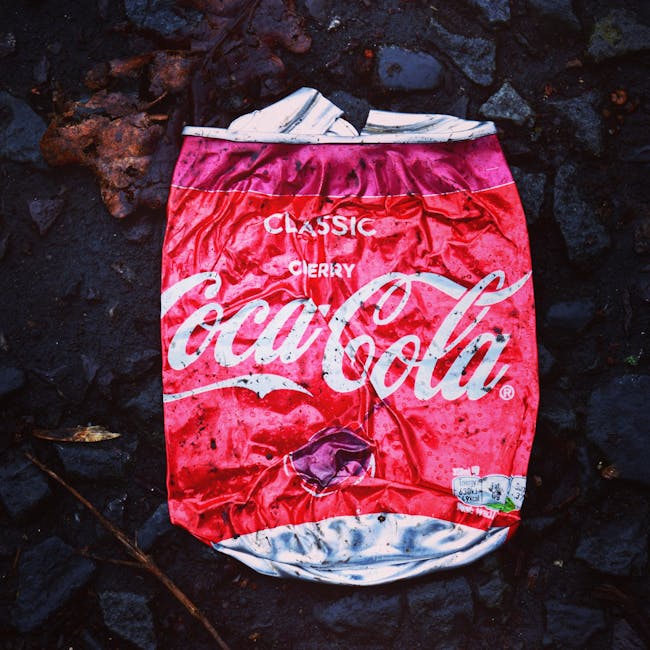A disposable paper cup with a white exterior and green printed text is lying on its side on a weathered brick surface, near a metal drainage grate embedded in the pavement. The brickwork appears dark 