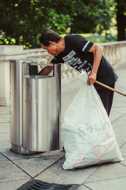 An outdoor scene showing a large pile of mixed household waste and recyclable materials, including cardboard boxes, plastic bags, paper piles, and discarded packaging, accumulated around multiple wast