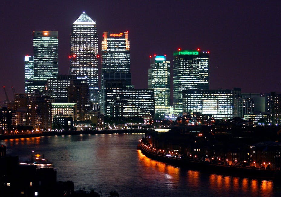 A modern urban scene featuring a tall, rectangular office building with a glass façade reflecting the sky and surrounding structures, situated behind a bridge spanning a body of water. The bridge is