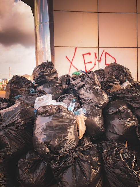 A worker wearing a blue shirt, black gloves, and a face mask is standing on the back of a large, open-top garbage truck in an urban setting during daytime. The truck is loaded with various household i